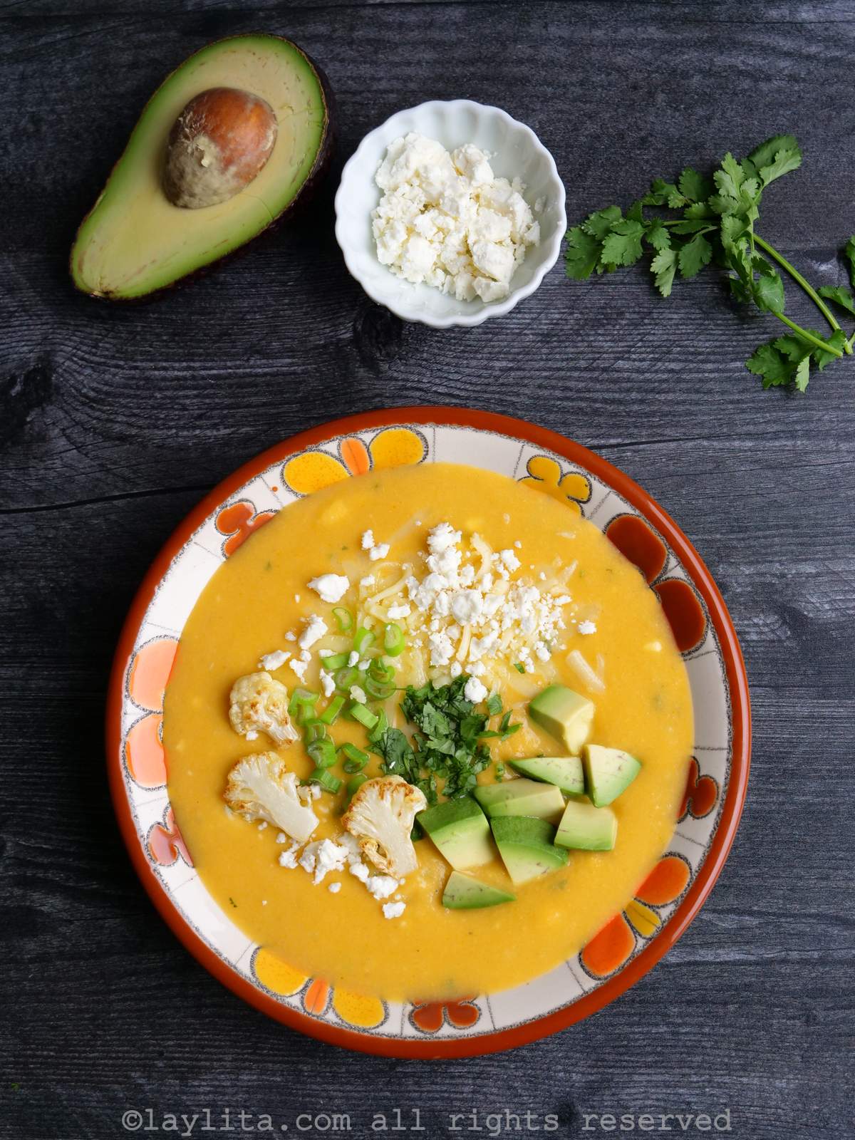 Uma tigela de sopa cremosa de couve-flor guarnecida com queijo e coentro picado, com mais queijo, abacate e coentro ao fundo.