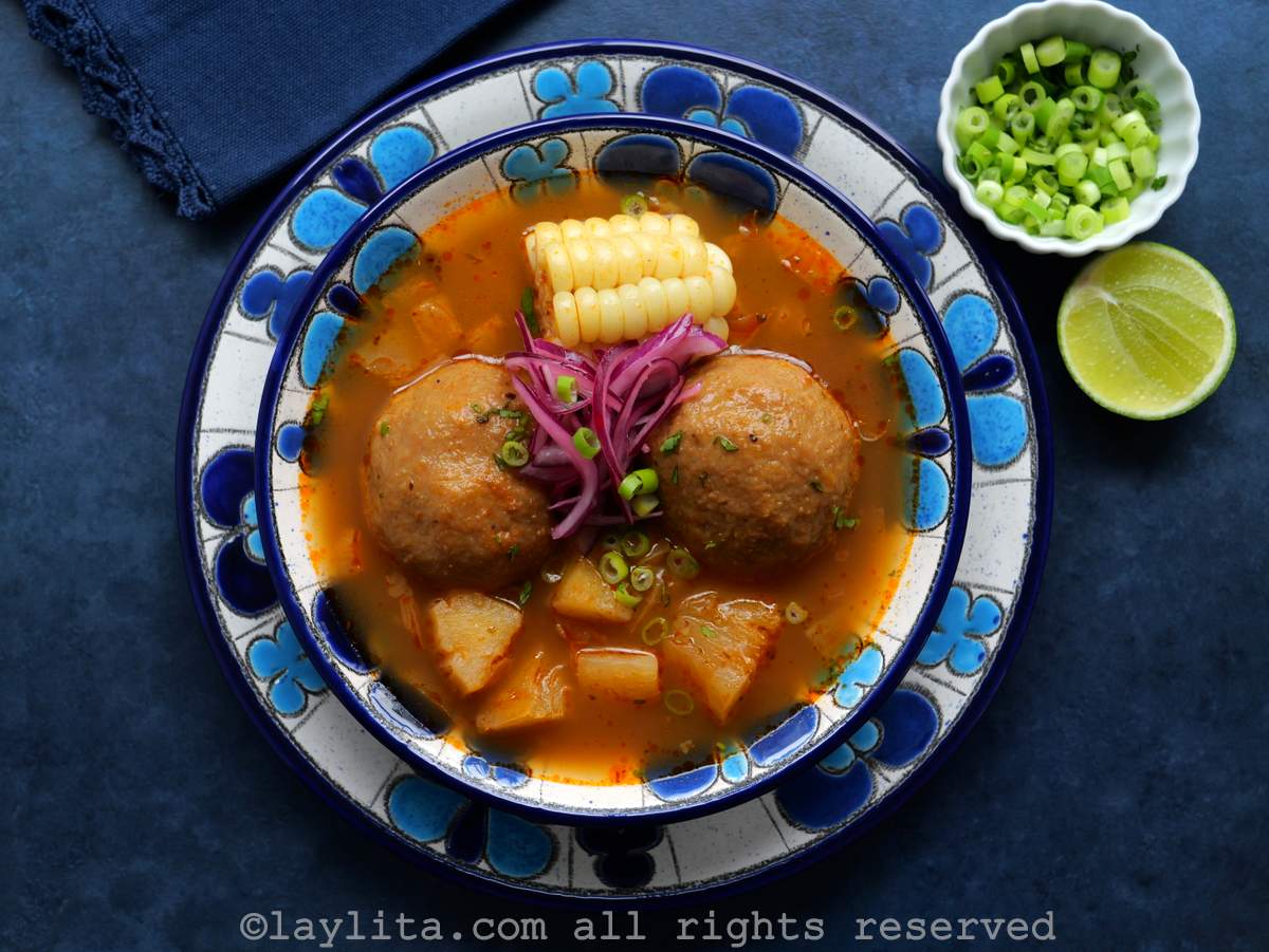 Vista superior de un plato sopero con caldo con dos bolas de plátano verde y un trozo de choclo, la sopa va decorada con cebollas curtidas