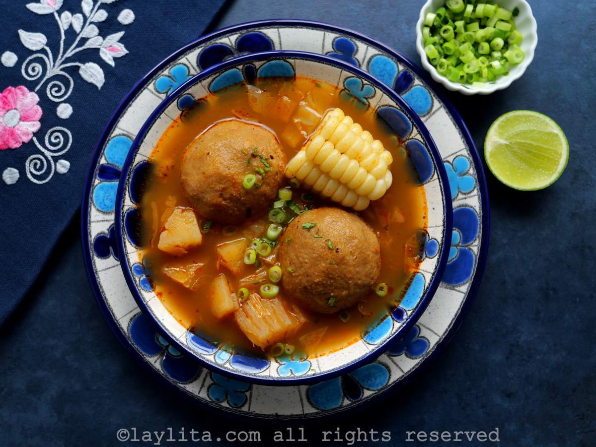Un plato sopero con flores azules relleno de caldo de bolas de verde, al lado hay una servilleta, una rodaja de limón y cebollín picado
