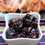 Un recipiente blanco lleno de salsa de ciruelas pasas encima de un mantel de colores, en el fondo se ven un plato con pavo asado cortado en rebanadas