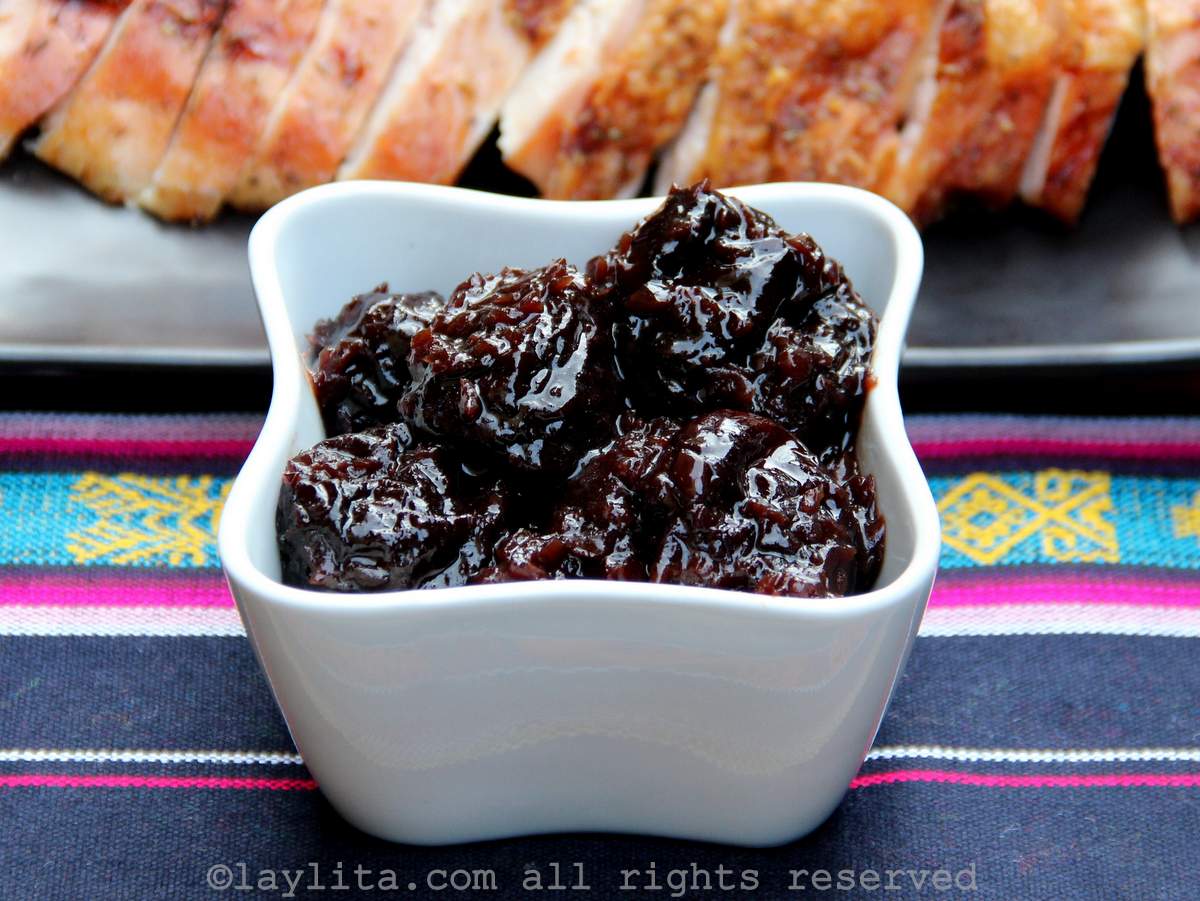 Salsa de ciruelas pasas en un recipiente blanco encima de un mantel de colores, en el fondo se ven un plato con pavo asado cortado en rebanadas