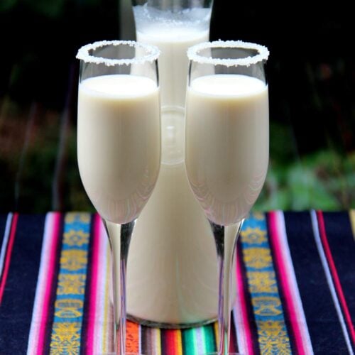 Dos copas de rompope con los bordes decorados de azucar y con una botella de rompope en el fondo