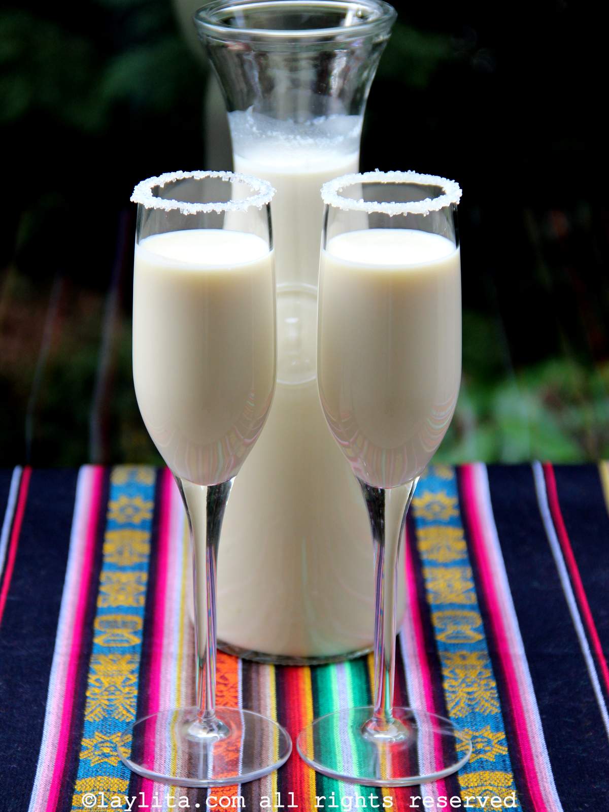 Dos copas de rompope con los bordes decorados de azucar y con una botella de rompope en el fondo