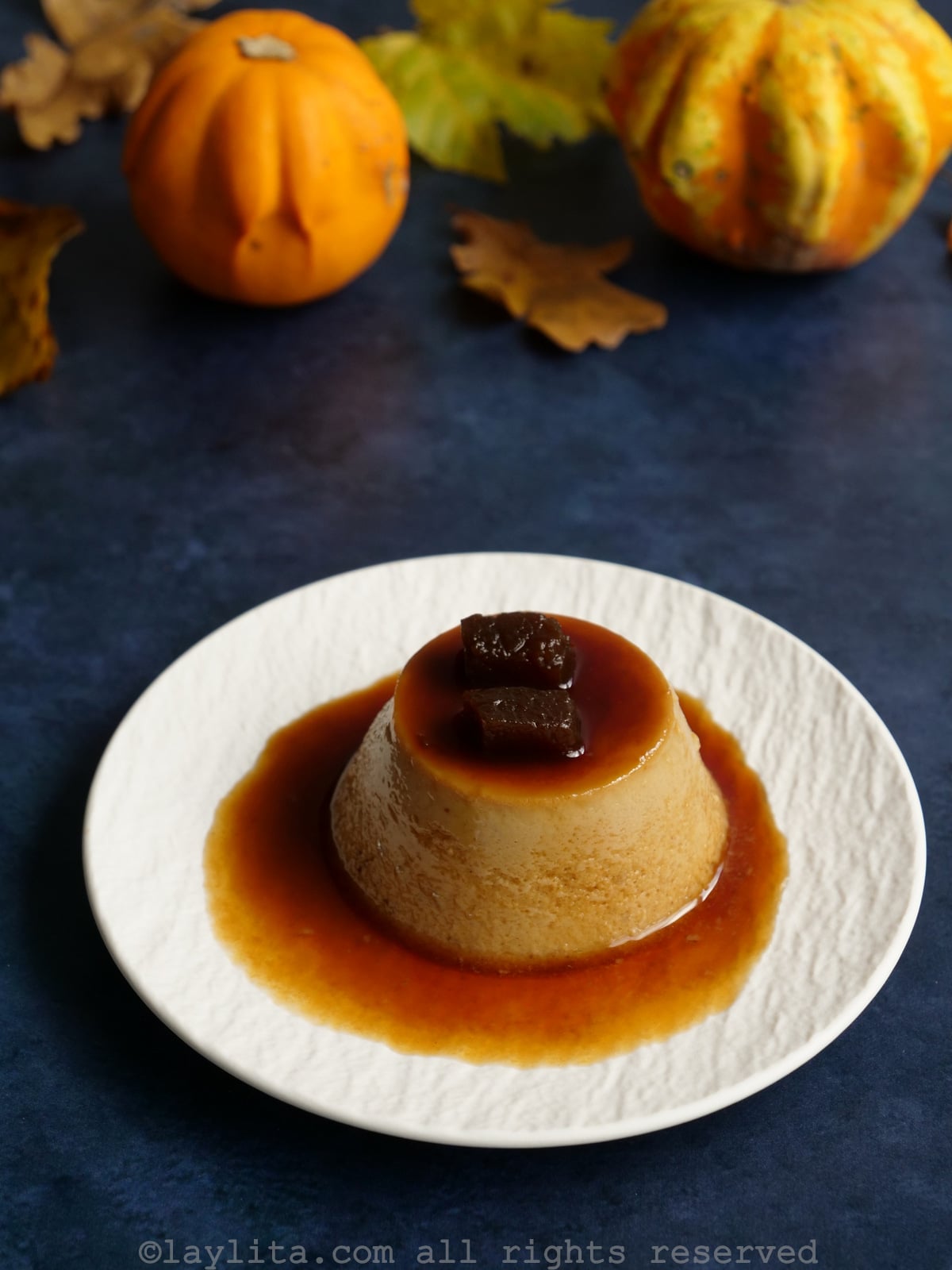 Un plato blanco con un flan de calabaza confitada y con calabacitas decorativas en el fondo