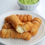 Varios tequeños de queso en un plato blanco con salsa de aguacate en el fondo