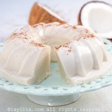 Un Tembleque Puertorriqueño con una rebanada faltante en un pedestal de tartas con un coco en el fondo
