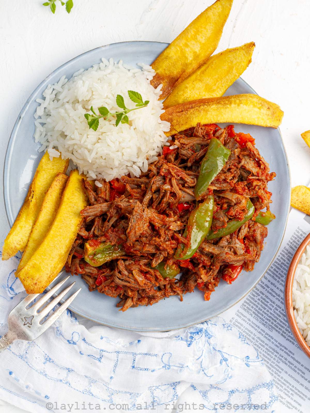 Ropa vieja cubana o carne desmenuzada guisada en un plato azul claro acompañado con arroz blanco y plátano frito