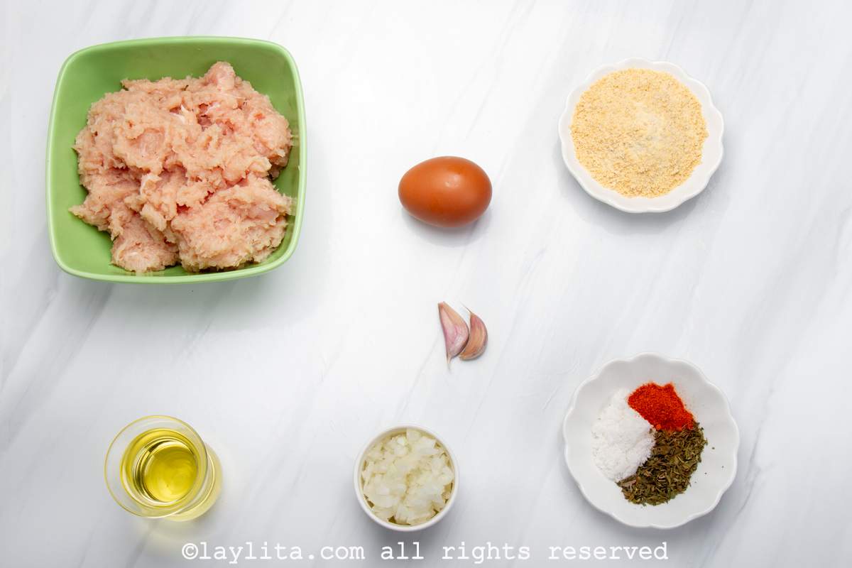 Ingredientes de las albóndigas de pollo en un mesa: Carne de pollo molida, huevo, migas de pan, aceite, ajos, cebolla picada, sal, pimentón dulce molido, y orégano