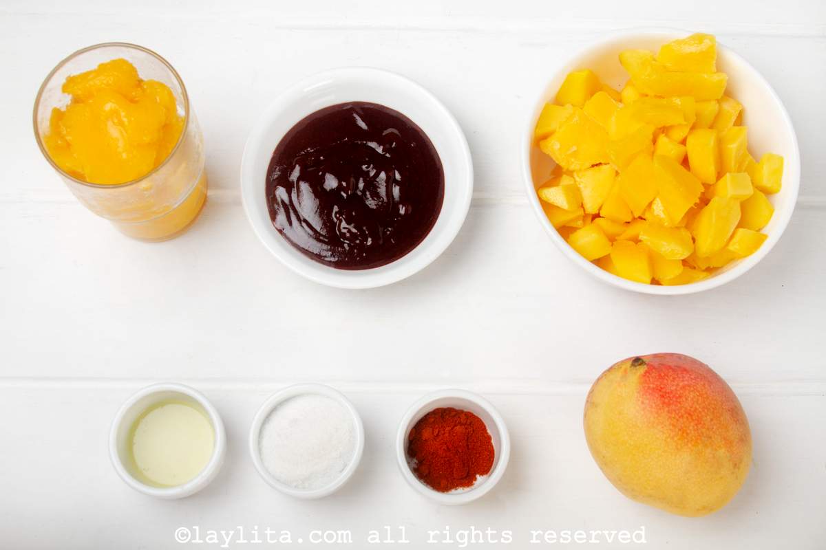 Ingredientes para preparar mangonadas en una mesa blanca: Mango congelado en cubos, jugo o néctar de mango, mango fresco, chamoy, chile en polvo, azucar, jugo de limón