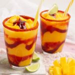 Dos vasos de vidrio llenos de mangonada, decorados con sorbete, limón, y mango fresco, con un fondo rosado