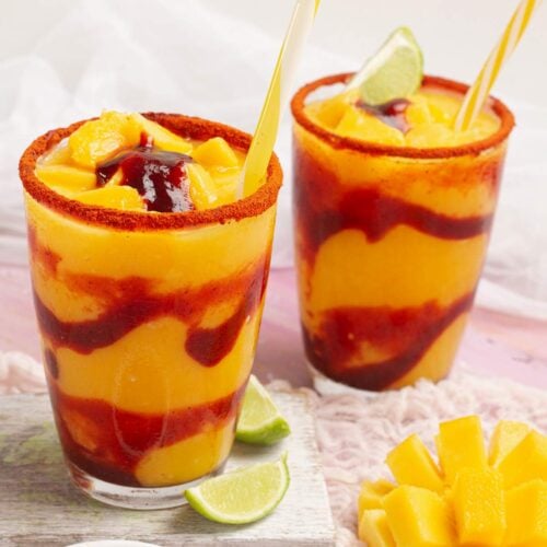 Dos vasos de vidrio llenos de mangonada, decorados con sorbete, limón, y mango fresco, con un fondo rosado