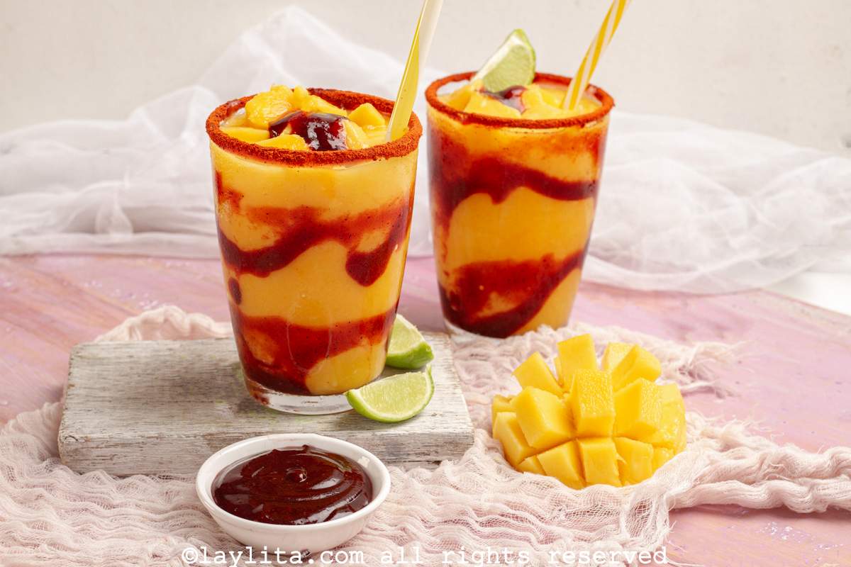 Dos vasos de vidrio llenos de mangonada, decorados con sorbete, limón, y mango fresco