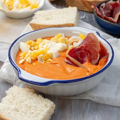 Un plato hondo blanco con borde azul lleno de salmorejo decorado con huevos duros y jamón serrano