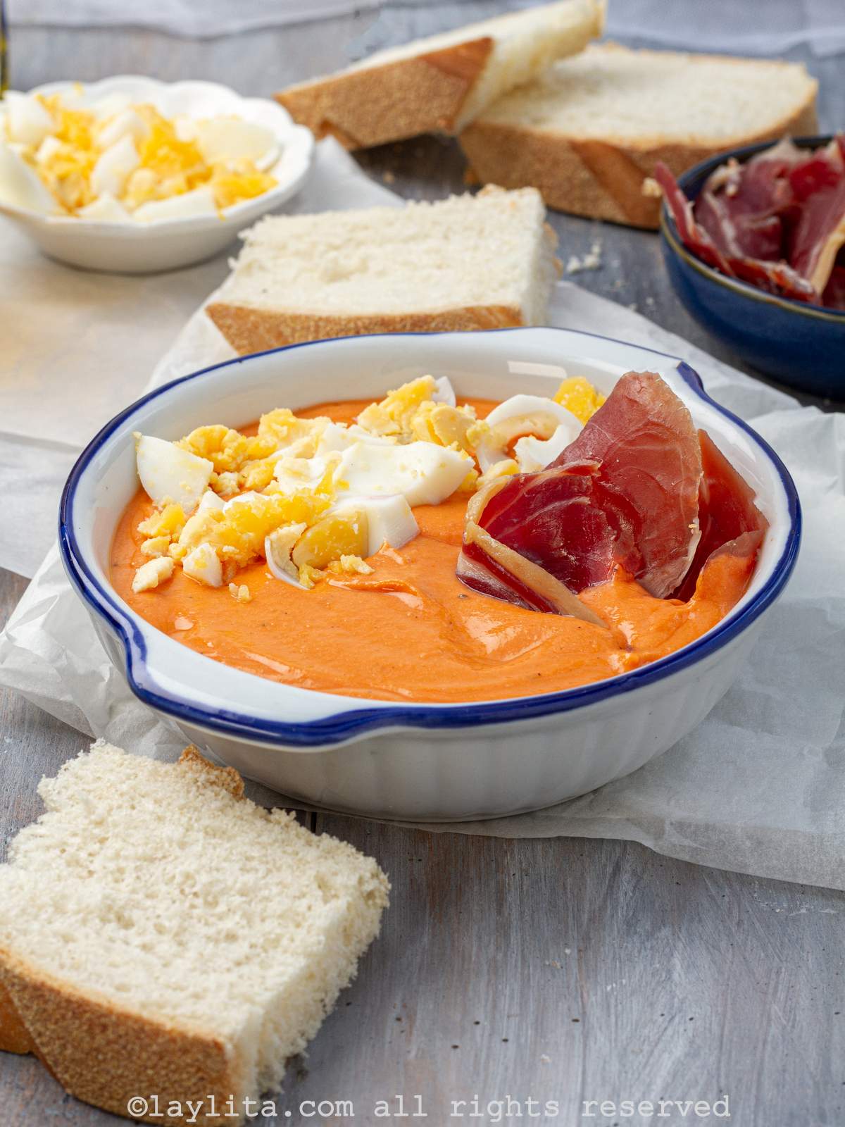 Un plato hondo blanco con borde azul lleno de salmorejo decorado con huevos duros y jamón serrano