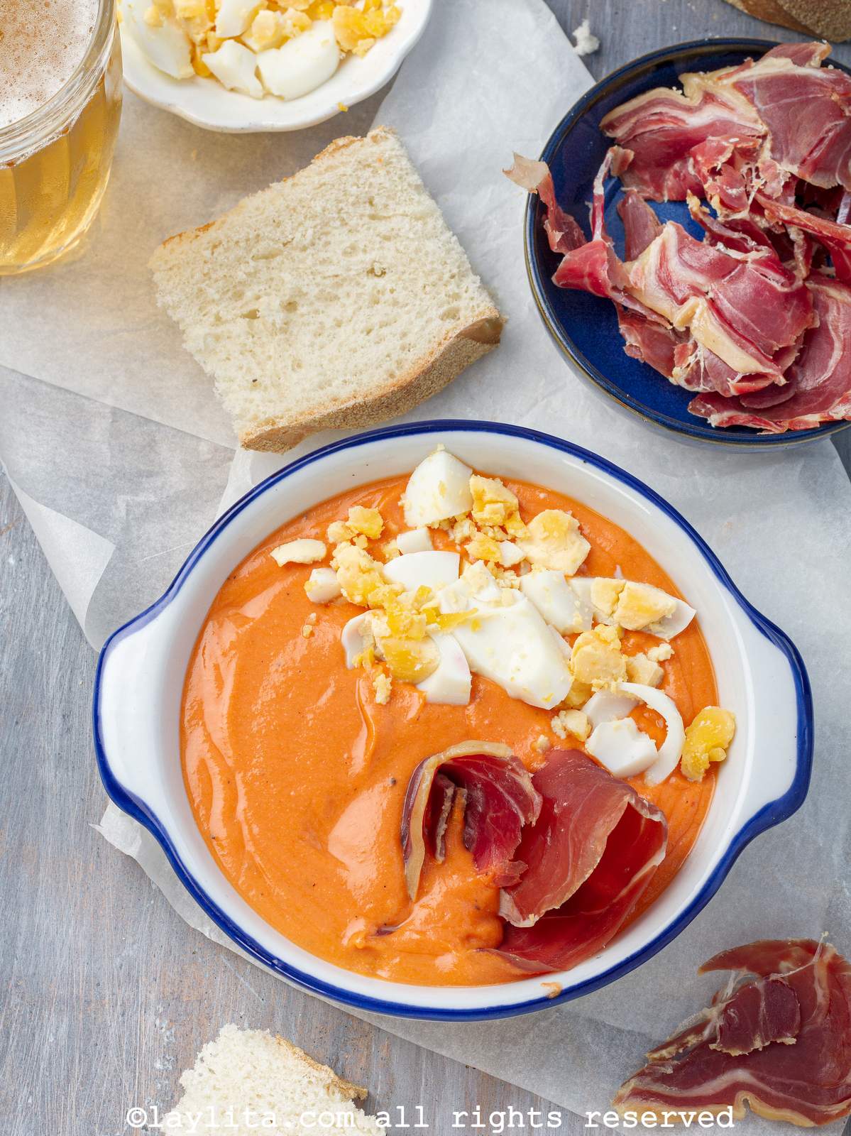 Un plato hondo blanco lleno de salmorejo decorado con huevos duros y jamón serrano. En el fondo se ven rodajas de pan, jamón serrrano y trozos de huevo duro
