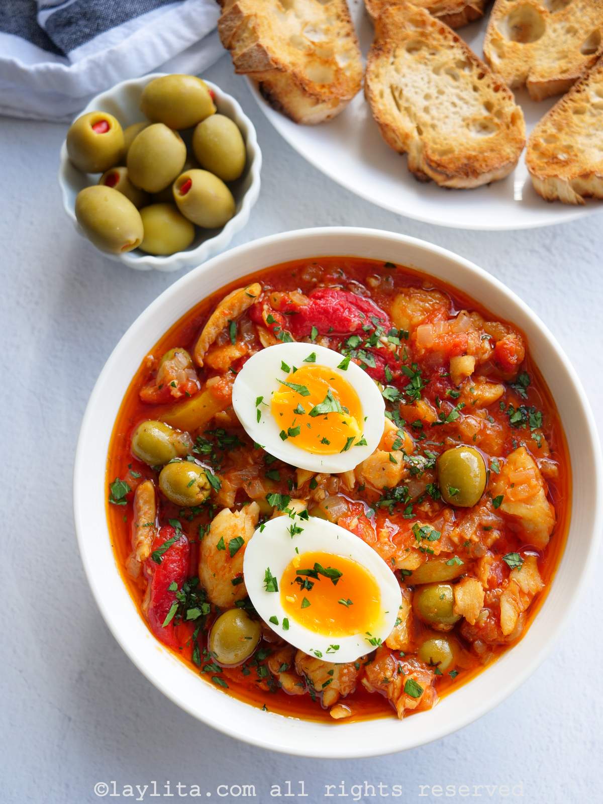 Un plato blanco con bacalao a la vizcaína , con aceitunas y pan en el fondo