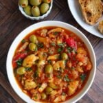 Un plato blanco con bacalao a la vizcaína , con aceitunas y pan en el fondo