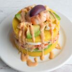 Causa peruana rellena con camarones en un plato blanco y decorada con camaron, aceituna y aguacate