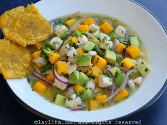 Un bol blanco lleno de ceviche de pescado con aguacate y mango, servido con dos patacones a un lado del bol