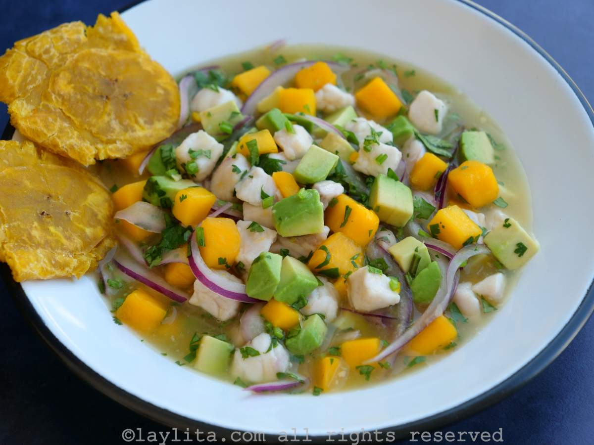 Un bol blanco lleno de ceviche de pescado con aguacate y mango, servido con dos patacones a un lado del bol