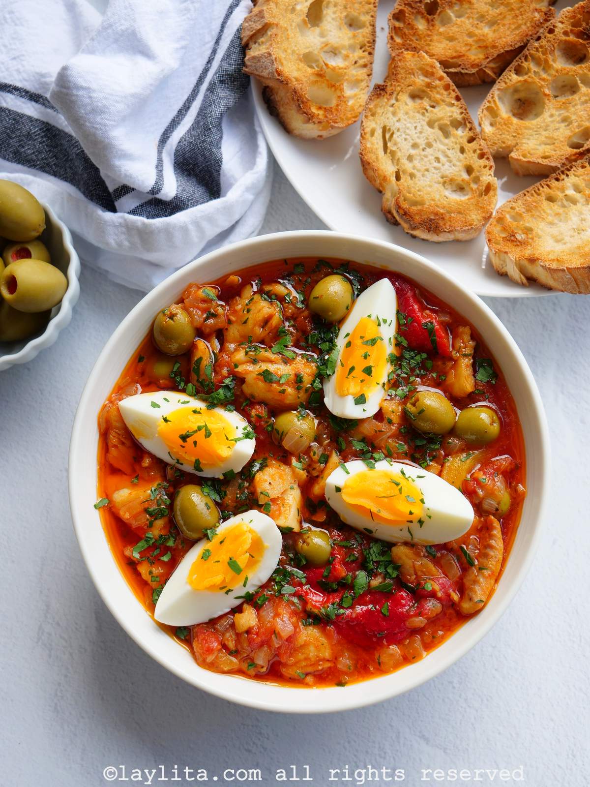 Un plato blanco con bacalao a la vizcaína , con aceitunas y pan en el fondo