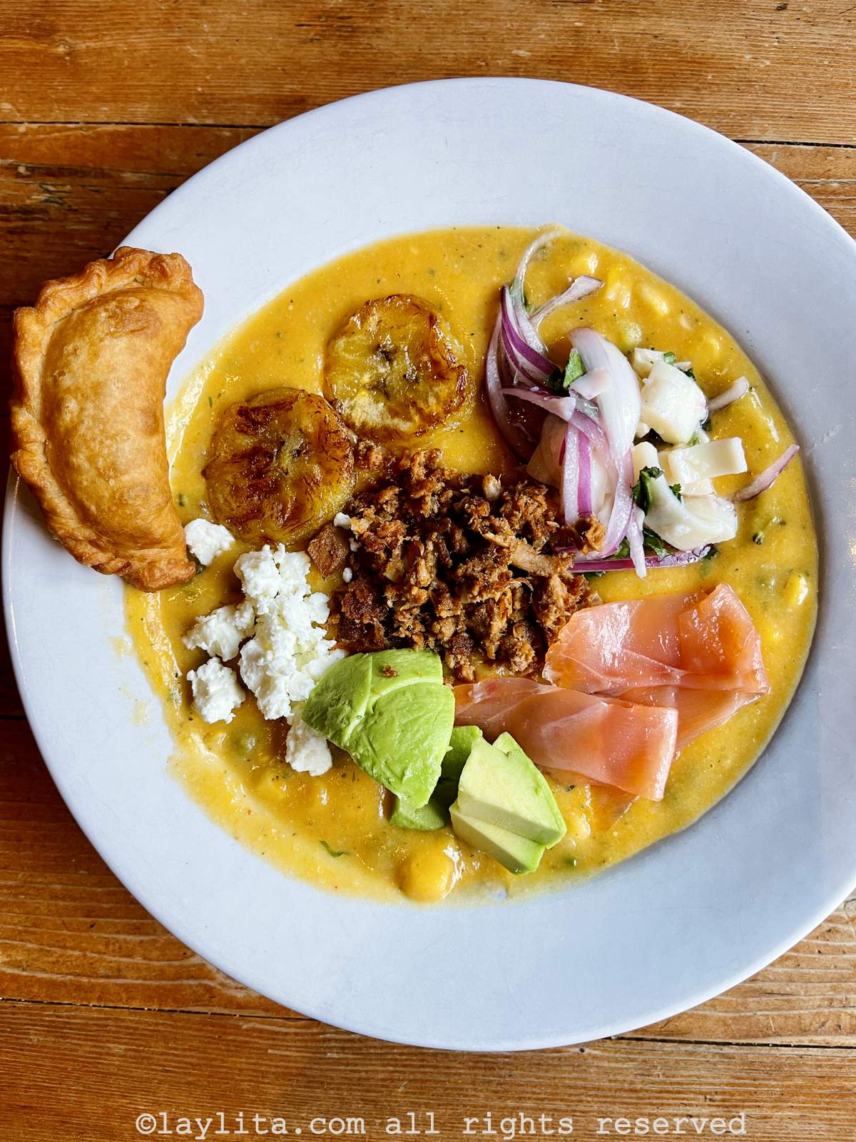 Un plato sopero blanco lleno de fanesca de atún adornado con aguacate, salmon, queso, maduros fritos, atun frito, ensalada de palmito