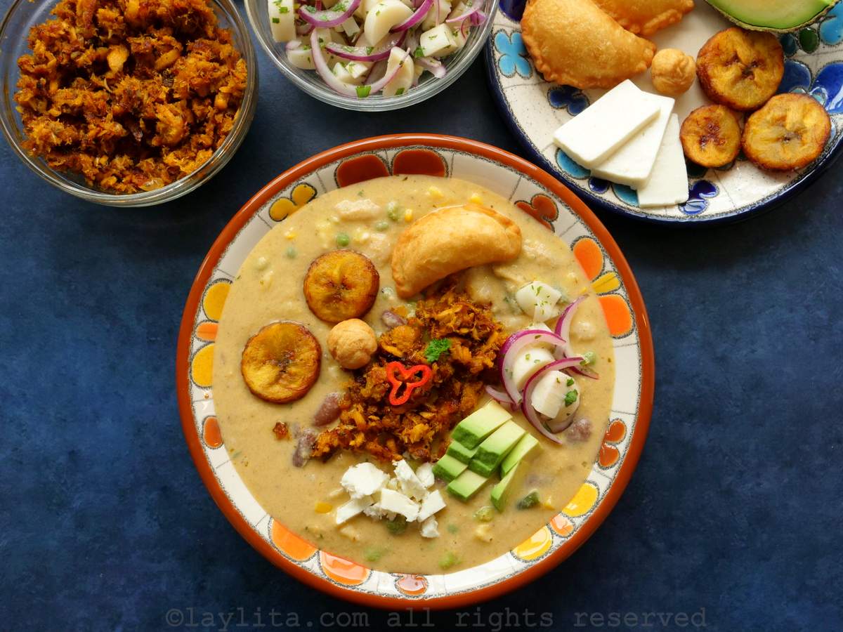 Un plato de sopa lleno de fanesca de atún adornado con aguacate, salmon, queso, maduros fritos, atun frito, ensalada de palmito, con guarniciones adicional al lado