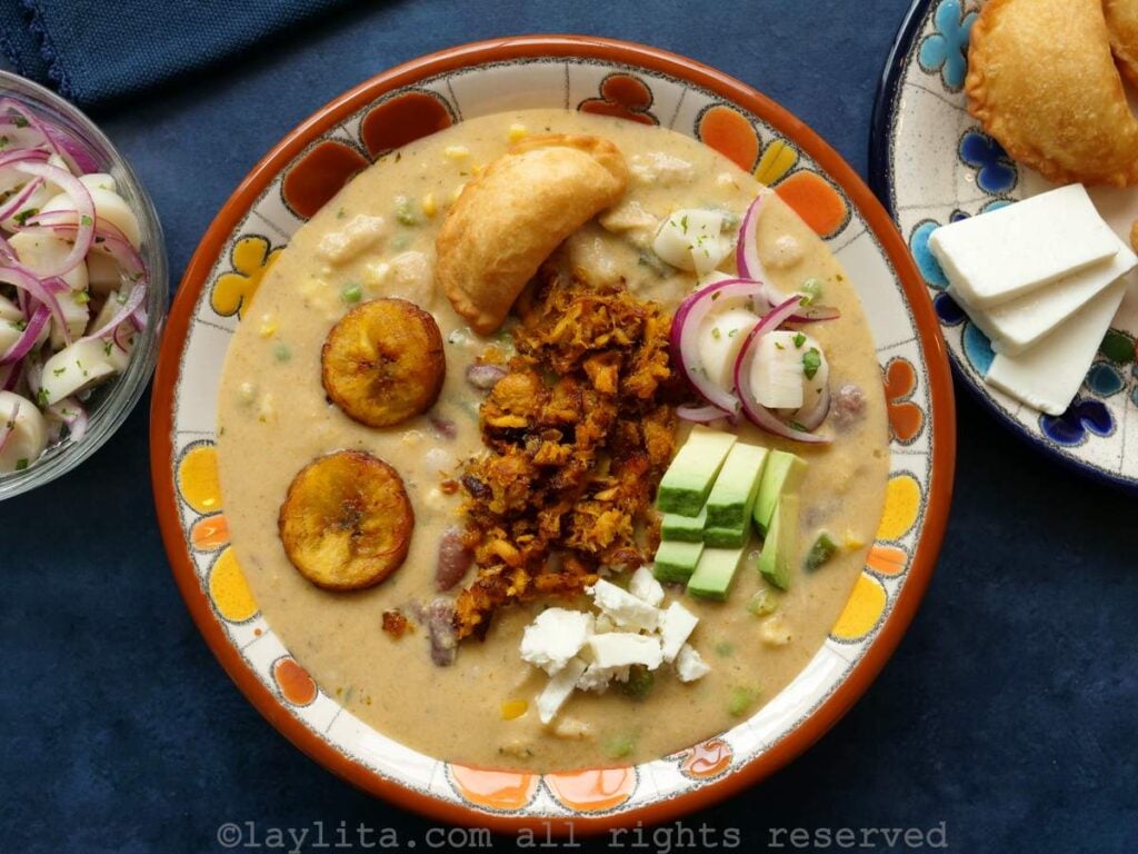 Un plato de sopa lleno de fanesca de atún adornado con aguacate, salmon, queso, maduros fritos, atun frito, ensalada de palmito, con guarniciones adicional al lado