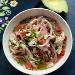 Un plato blanco lleno de ceviche de pollo con una mitad de aguacate al lado