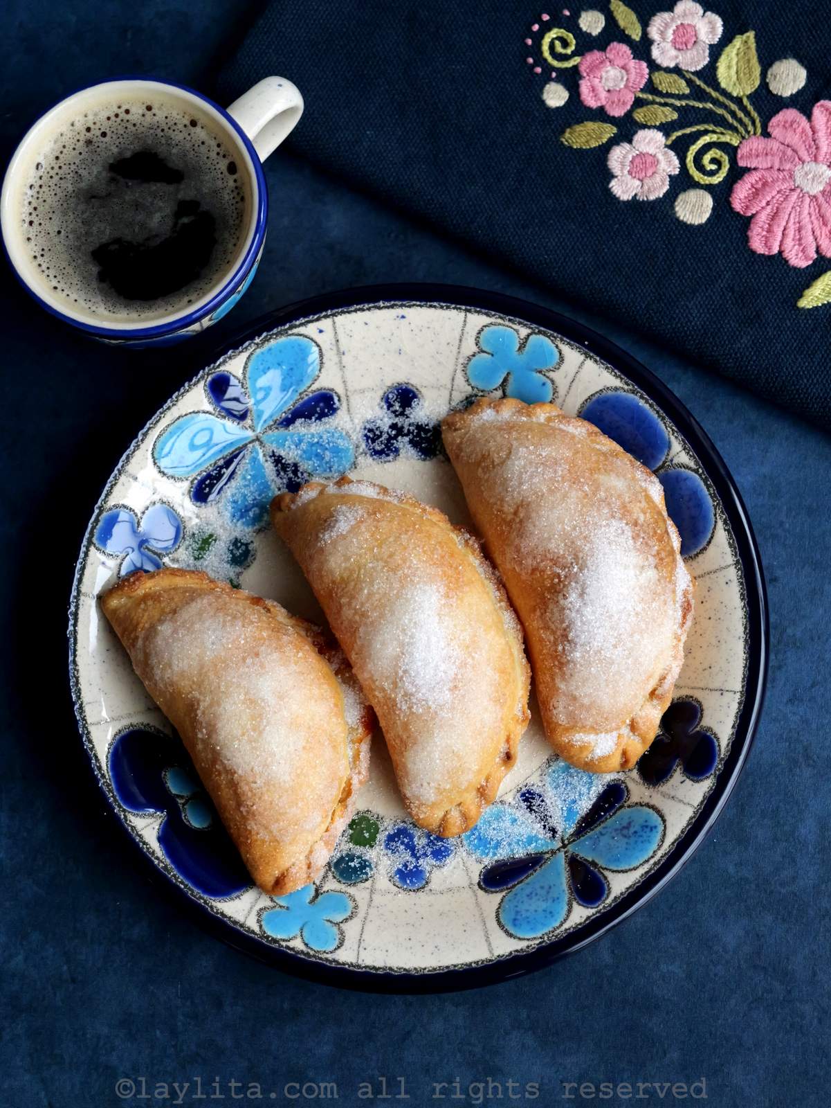 Tres empanadas de queso con azúcar en un plato floreado con una taza de café y una servilleta bordada al lado