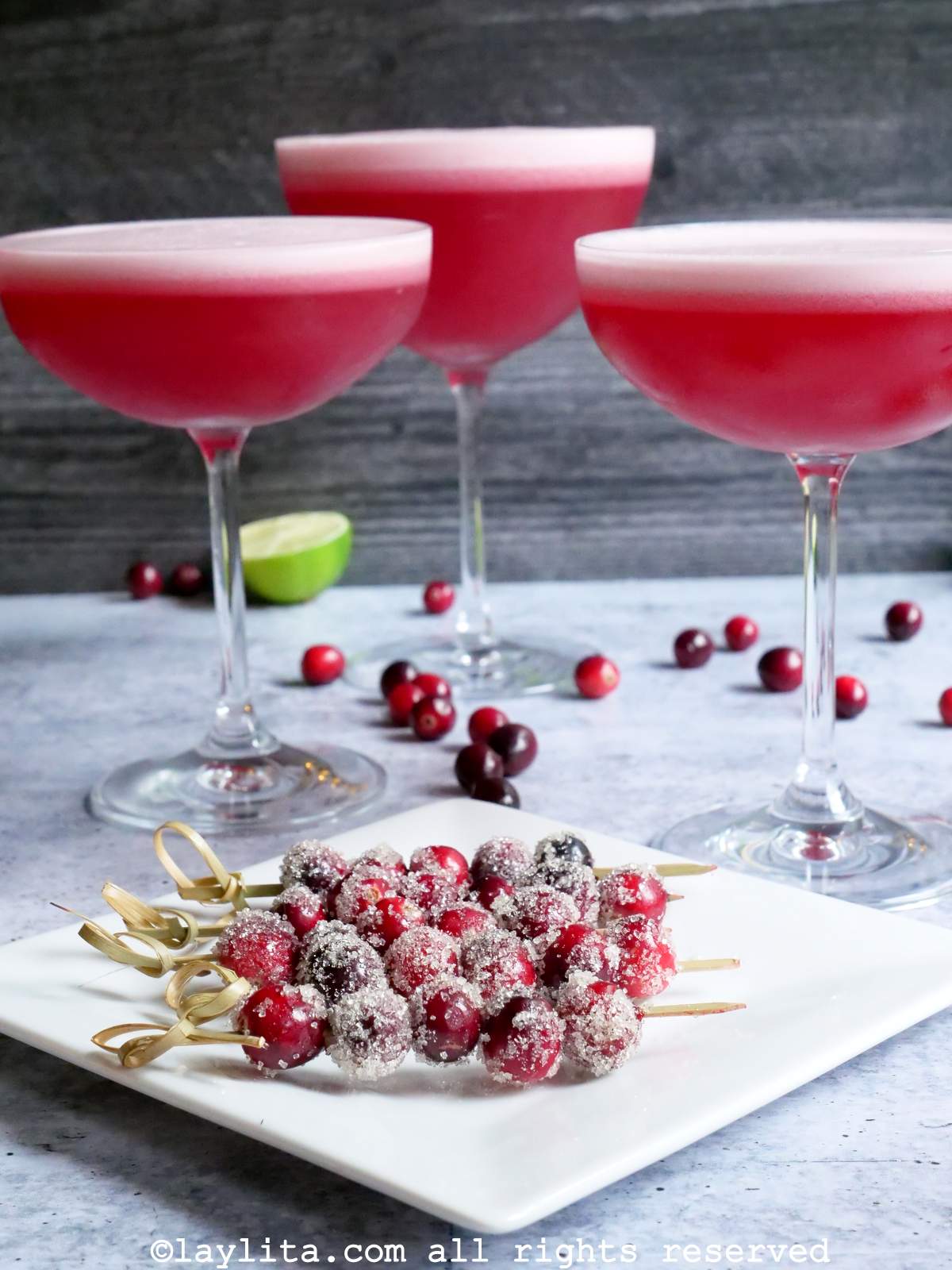 Arándanos azucarados en palillos en un plato blanco, en el fondo se ven tres copas llenas de pisco sour de arándano rojo