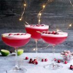 Tres copas llenas de pisco sour de arándano rojo, decorados con arándanos, en el fondo se ven mas arandanos , limones y luces