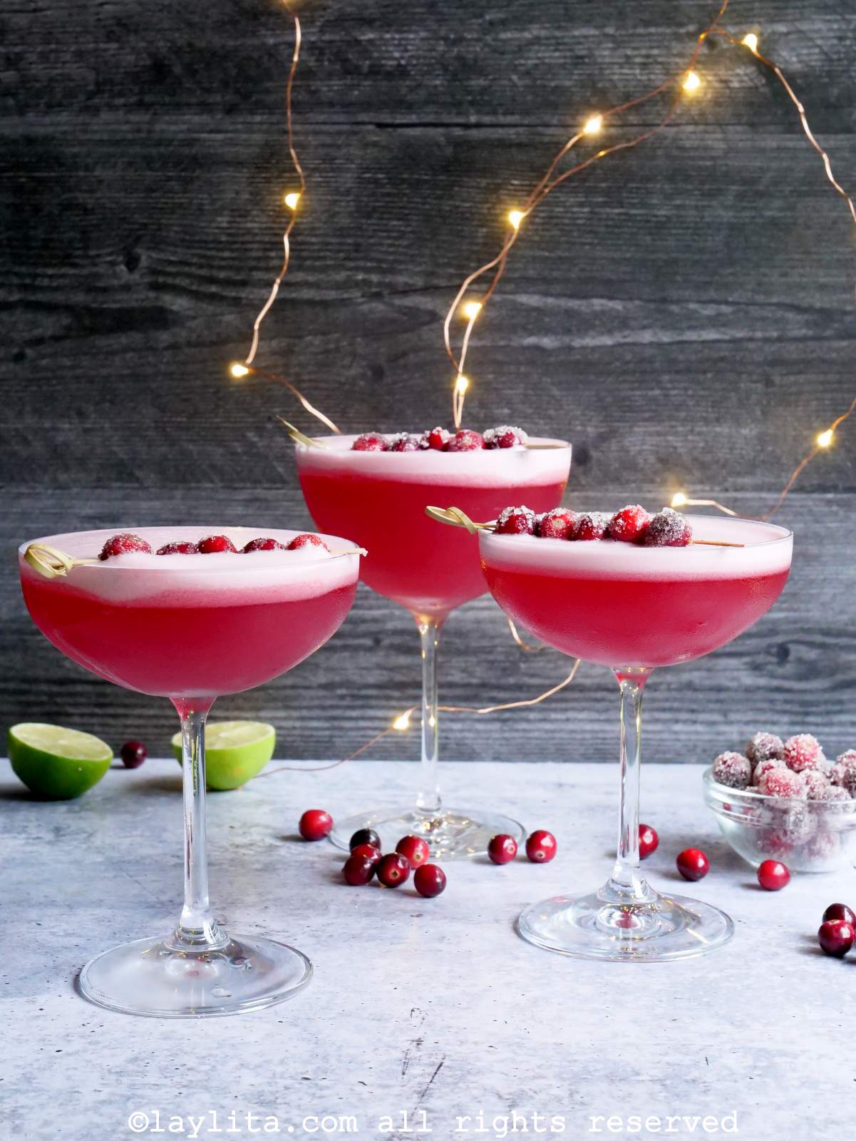Tres copas llenas de pisco sour de arándano rojo, decorados con arándanos, en el fondo se ven mas arandanos , limones y luces
