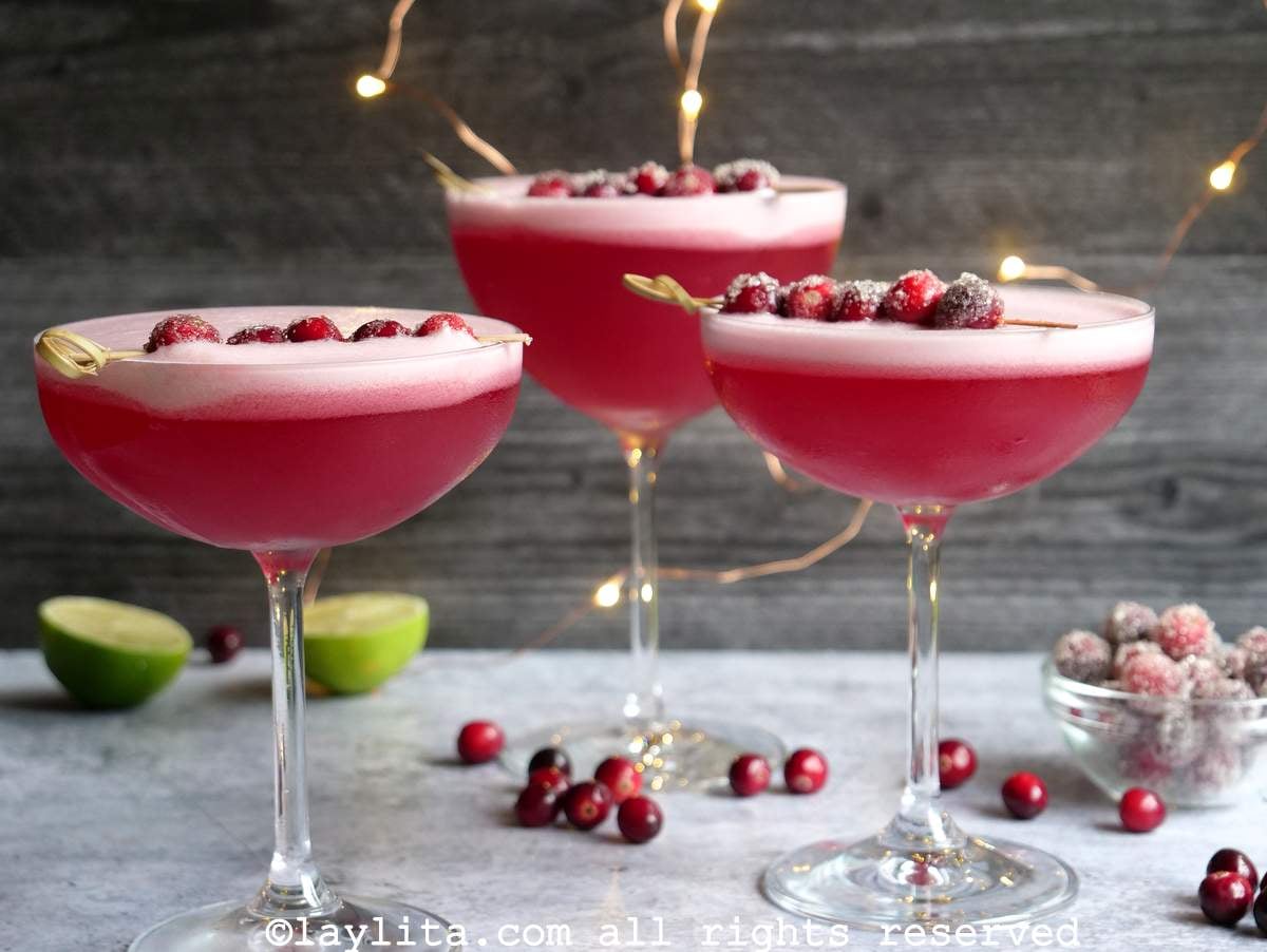 Tres copas llenas de pisco sour de arándano rojo, decorados con arándanos y luces en el fondo