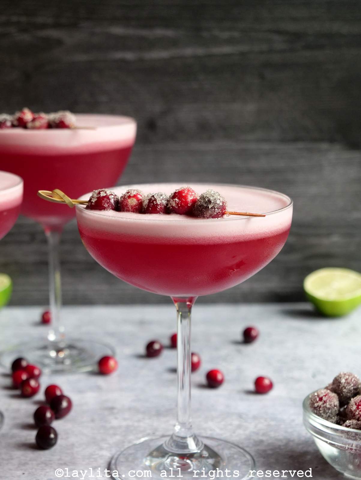 Cóctel pisco sour de arándano rojo en una copa con dos otras copas en el fondo, tambien se ven arandanos rojos y un limon cortado por la mitada en el fondon