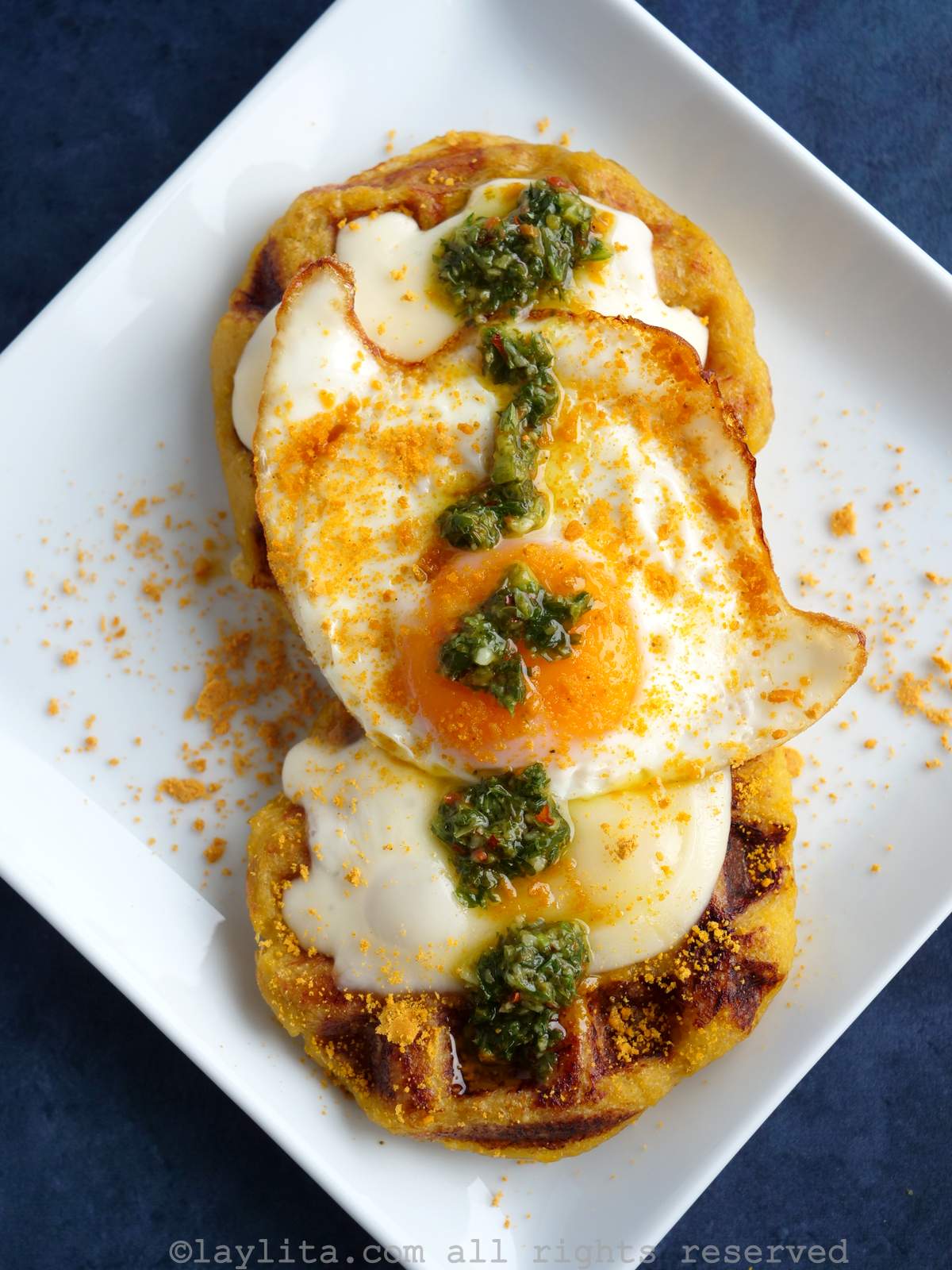 Un plato blanco con dos gofres de plátano macho cubiertos con queso, huevo frito y salsa chimichurri