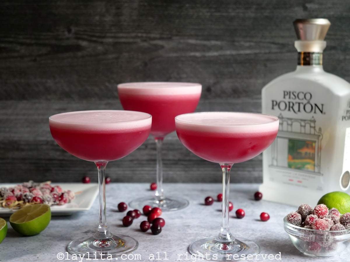 Tres copas llenas de pisco sour de arándano rojo, con una botella de pisco en el fondo y frutas alrededor de las copas
