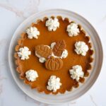 Un plato blanco pay de calabaza con base de galletas y decorado con crema batida y galletas