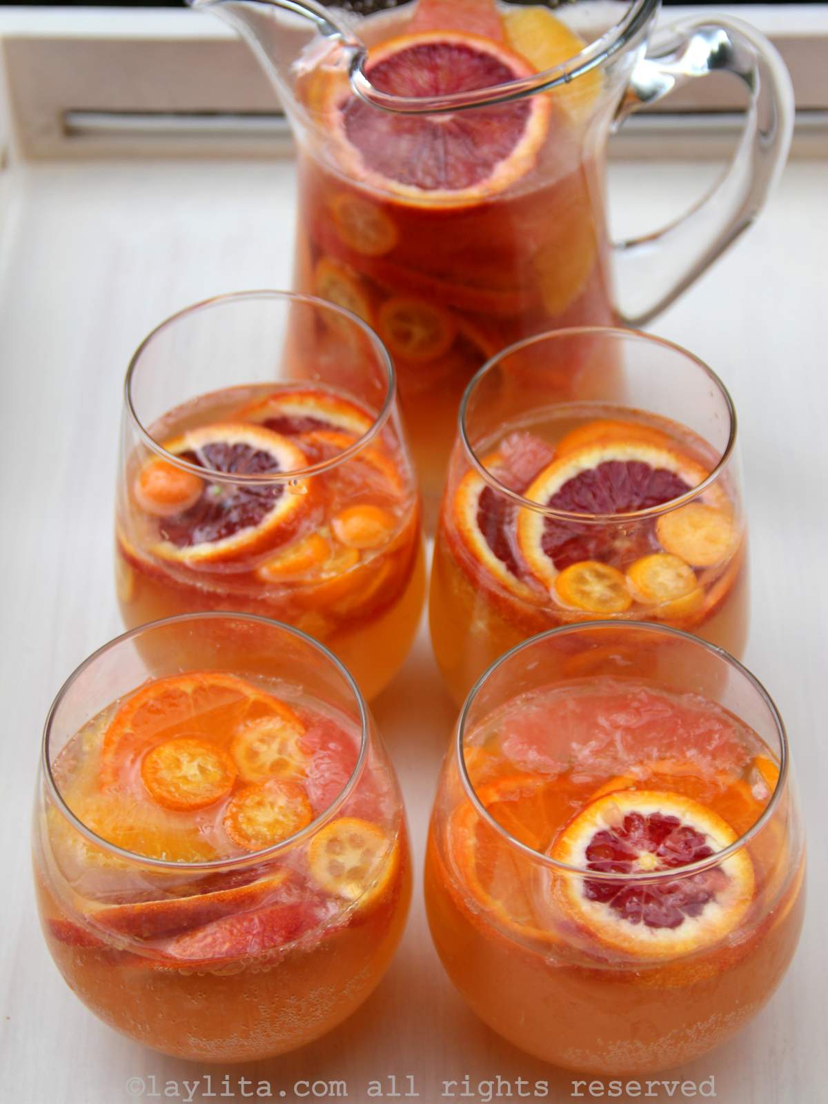 Una bandeja blanca con una jarra y cuatros vasos llenos de sangría de vino moscatel con rodajas de frutas cítricas