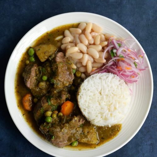 Un plato blanco con seco de res peruano, arroz blanco, fréjoles y salsa criolla, en una superficie azulada