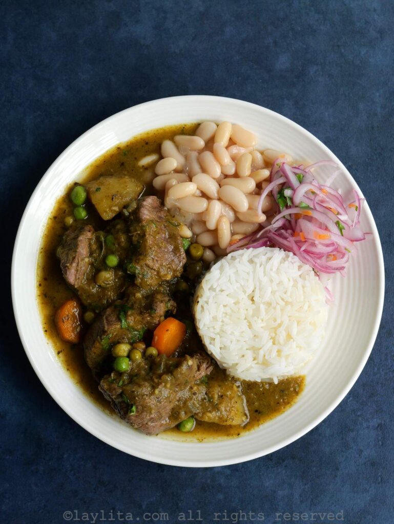 Un plato blanco con seco de res peruano, arroz blanco, fréjoles y salsa criolla, en una superficie azulada