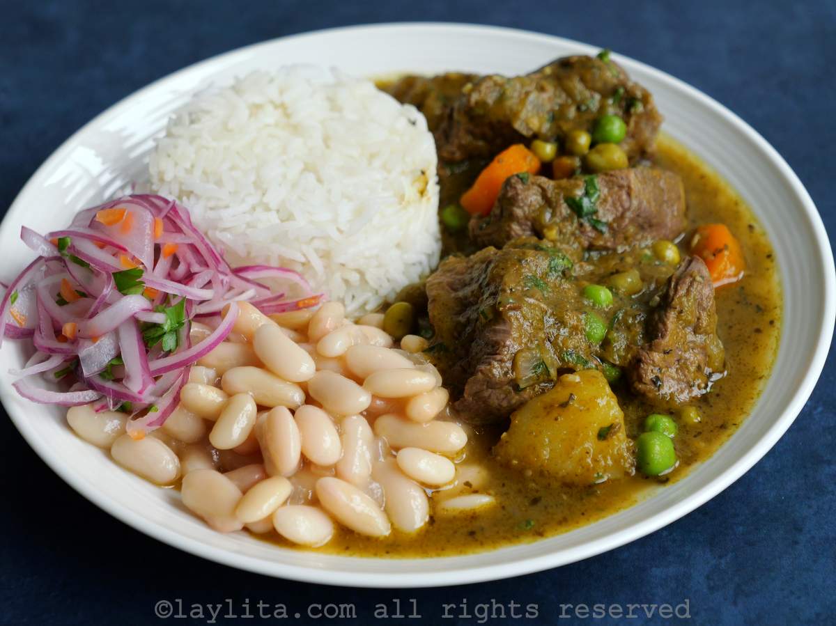 Seco de carne peruano en un plato blanco, acompañado con arroz, frejoles y salsa criolla de cebolla