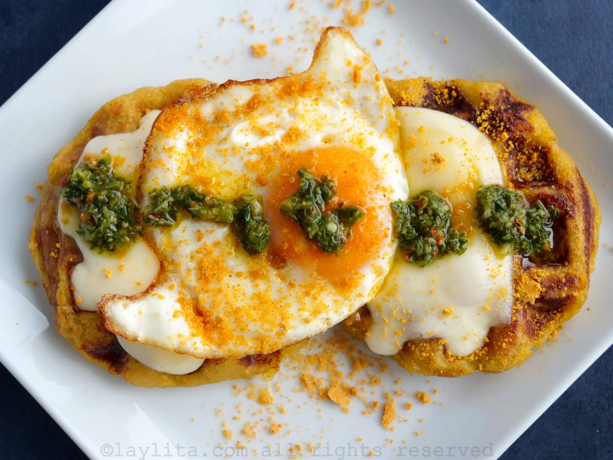 Un plato blanco con dos gofres de plátano con queso, huevo frito y chimichurri por encima