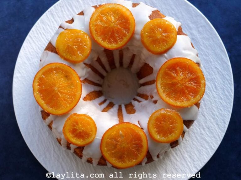 Una torta de naranja decorada con glaseado y rodajas de naranja confitadas en un plato blanco en una superficie azulada