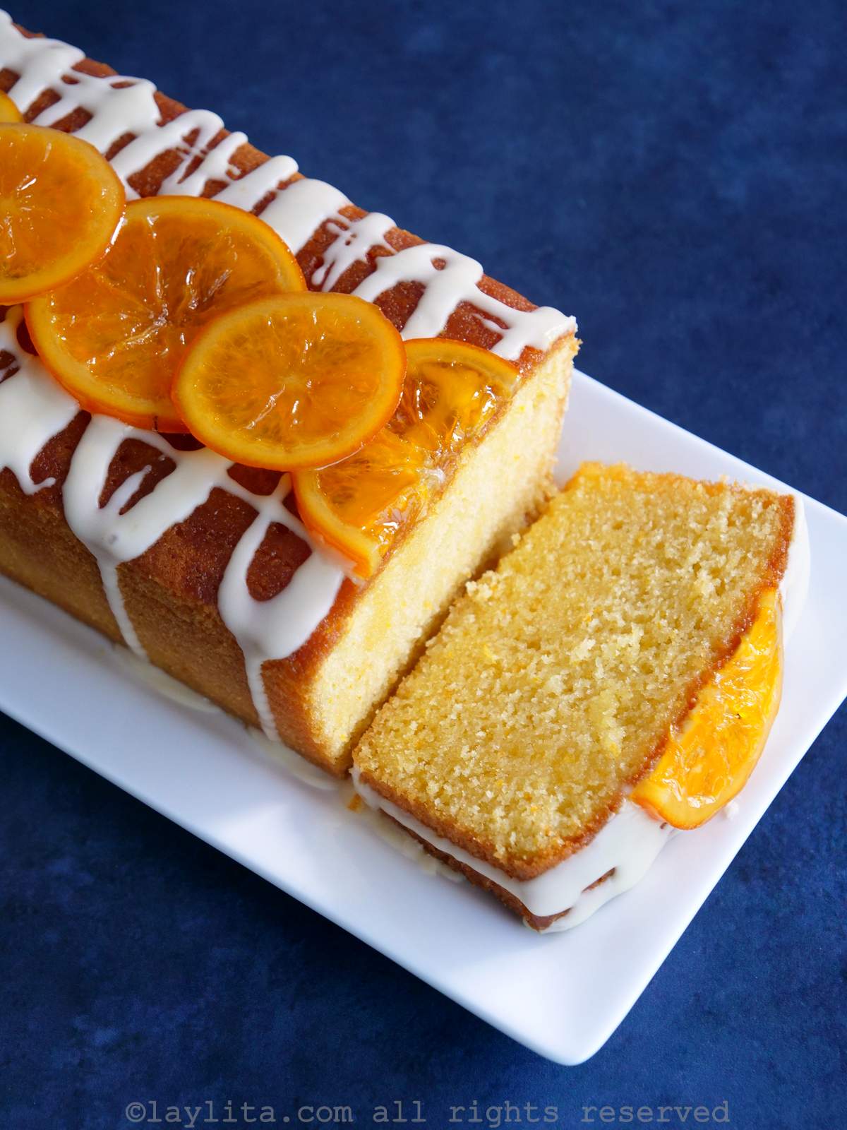 Un bizcocho de naranja decorado con naranjas confitadas con una rebanada cortada en un plato blanco