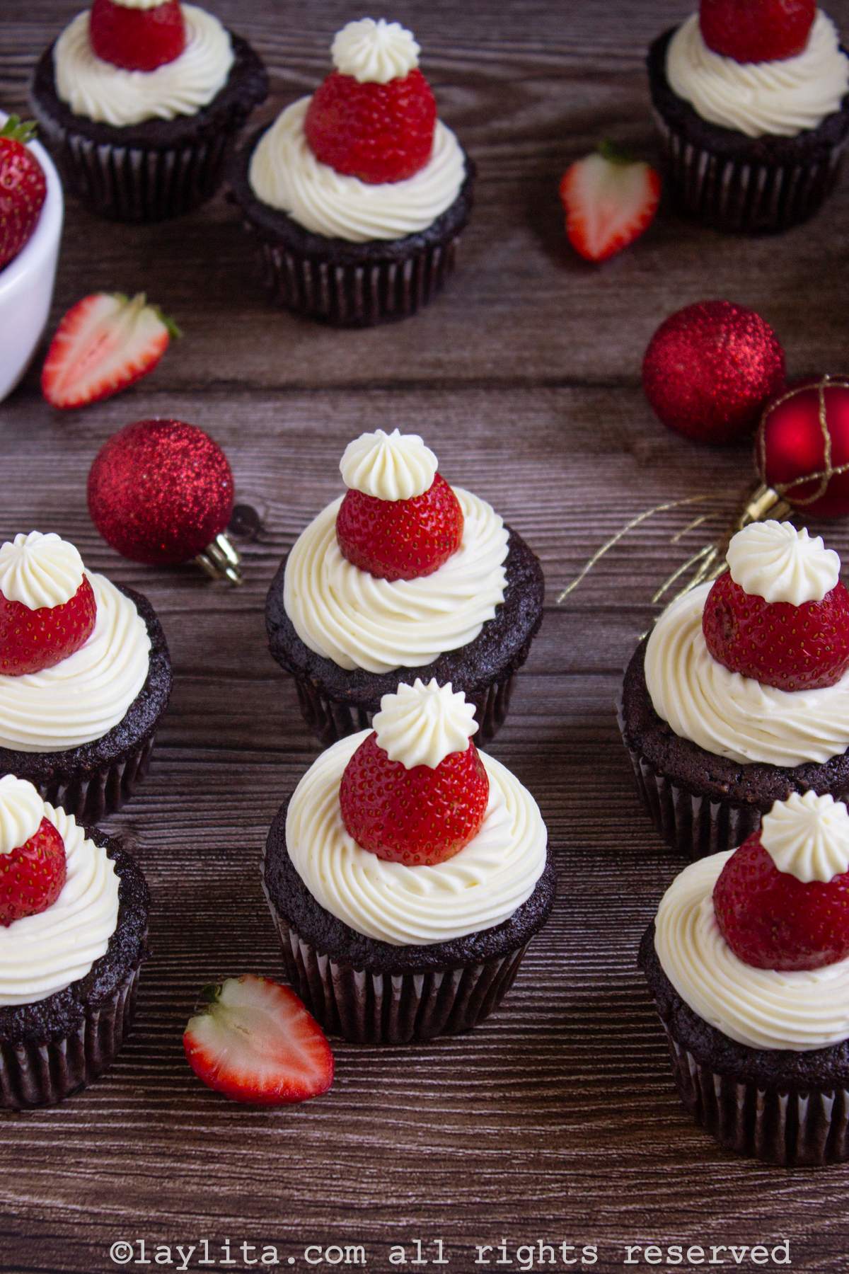 Varios cupcakes navideños de Papá Noel en una mesa de madera con fresas y bombillos rojos de Navidad