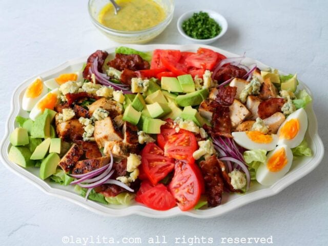 Una fuente llena de ensalada de pollo con verduras, en la parte de atras hay un bol con vinagreta y otro bol con perejil picado