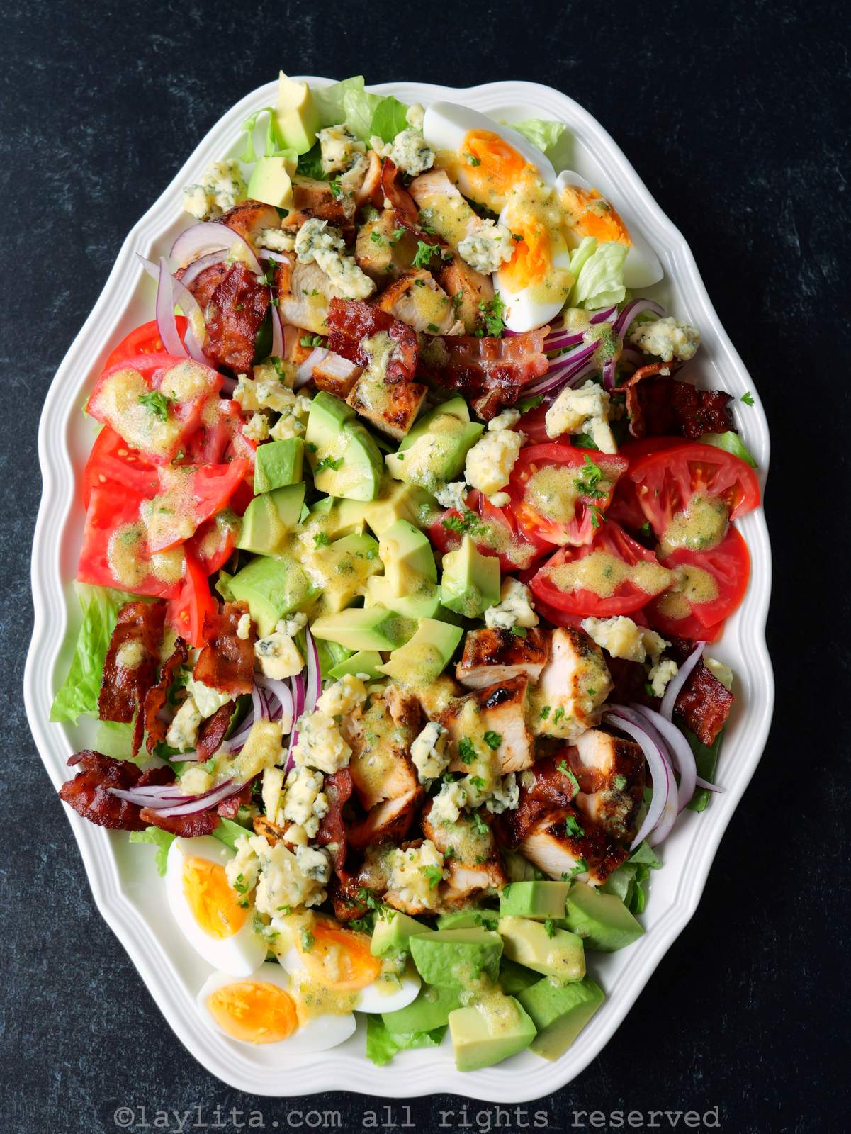 Una fuente blanca llena de ensalada de pollo con aguacate, tomate, huevo duro, cebolla, tocino, lechuga y aderezo de mostaza