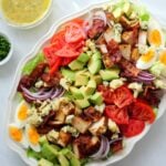 Una fuente ovalada blanca llena de ensalada con pollo, aguacate, tomate, huevos, cebolla, tocino y queso con un bol de vinagreta al lado