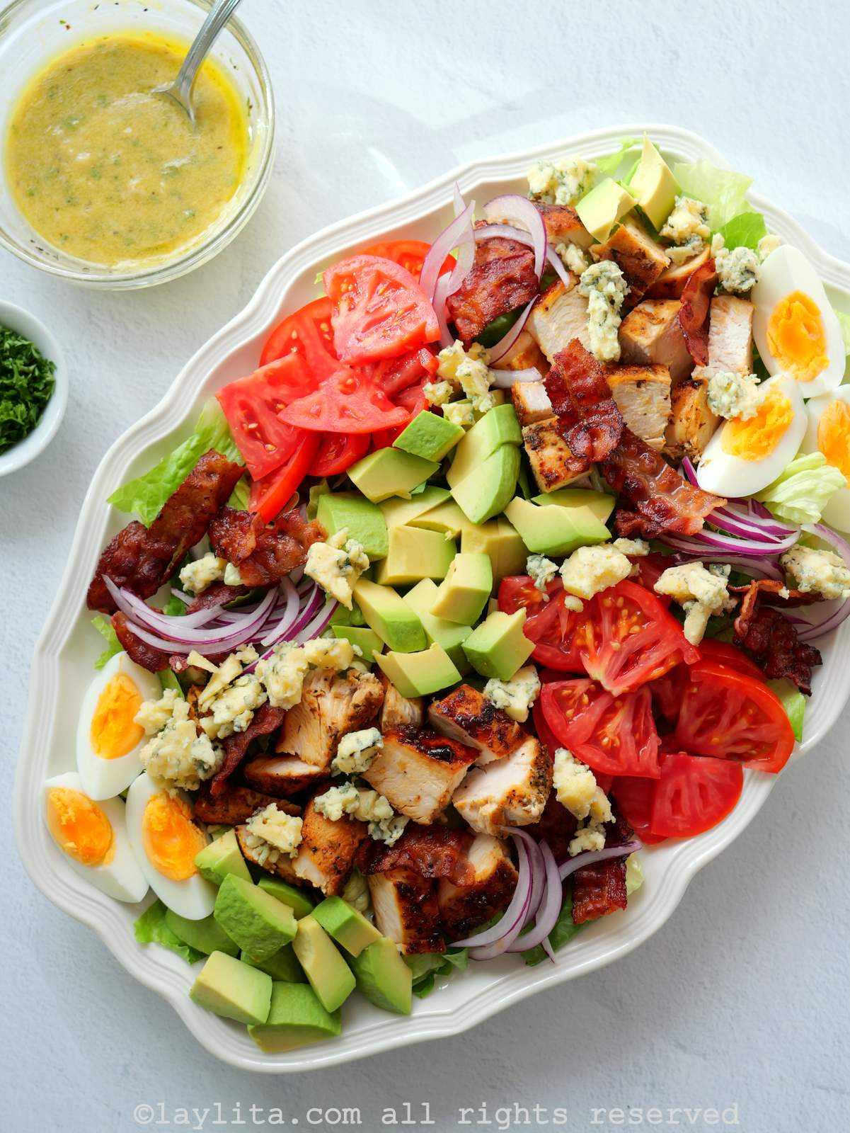 Una fuente ovalada blanca llena de ensalada con pollo, aguacate, tomate, huevos, cebolla, tocino y queso con un bol de vinagreta al lado